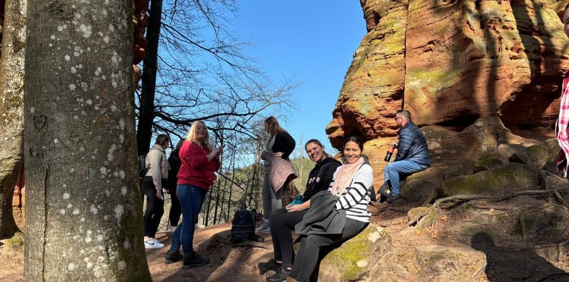Bunte Gruppe von Menschen beim Wandern in den Felsen des Pfälzerwaldes an einem sonnigen Tag. Zwei Frauen sitzen entspannt auf einem Felsen, während andere unterwegs sind. Der Himmel ist klar und blau, umgeben von Bäumen und festem Sandboden.