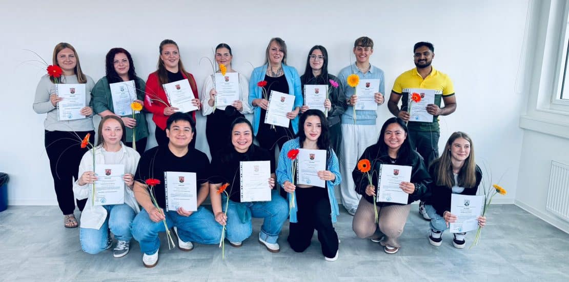 Erwachsene und Jugendliche mit Abschlusszeugnissen und Blumen bei Bildungszentrum Pirmasens.